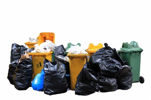 A collection of various rubbish bins and bags positioned on a paved surface outdoors, including four wheelie bins in green, yellow, and orange with closed lids and visible textures, alongside numerous black plastic rubbish bags that appear to be full and tied at the top, some leaning against the bins. The scene shows the waste in a mixed arrangement, with a blue bag among the black bags. The background is plain white, emphasizing the waste containers and rubbish bags. This setup reflects typical waste collection and rubbish removal practices, likely in a residential or commercial area within Richmond upon Thames, aligning with the services offered by Garden Clearance Richmond upon Thames for rubbish removal needs.