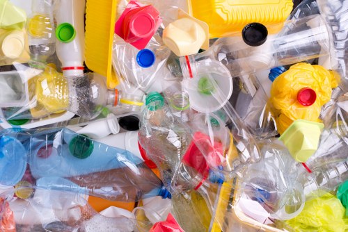 A large collection of crushed and uncrushed plastic bottles, predominantly transparent with coloured caps in red, green, blue, yellow, and black, scattered randomly in a pile on a flat surface. The bottles vary in size and shape, some with labels still attached, while others are plain. Visible in the background are fragments of plastic packaging, caps, and bottle necks, indicating a mix of waste materials. The scene is brightly lit, emphasizing the reflective surfaces and translucent qualities of the plastics. This image illustrates typical rubbish associated with plastic waste collection and disposal, relevant to rubbish removal services offered by companies like Garden Clearance Richmond upon Thames, particularly in areas like Richmond upon Thames or nearby postcode districts. The environment appears to be an indoor or sheltered area designated for waste sorting or accumulation, highlighting the accumulation of plastic waste awaiting removal. Such detailed visuals support local waste management activities and emphasize the importance of proper rubbish disposal and recycling efforts within the community.