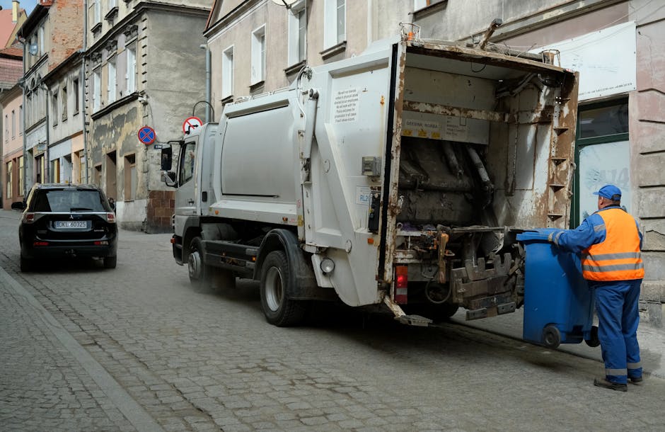 A small, dark red flatbed truck positioned on an urban street in front of a modern, multi-storey office building with large glass windows. The truck's cargo bed is filled with various types of waste m